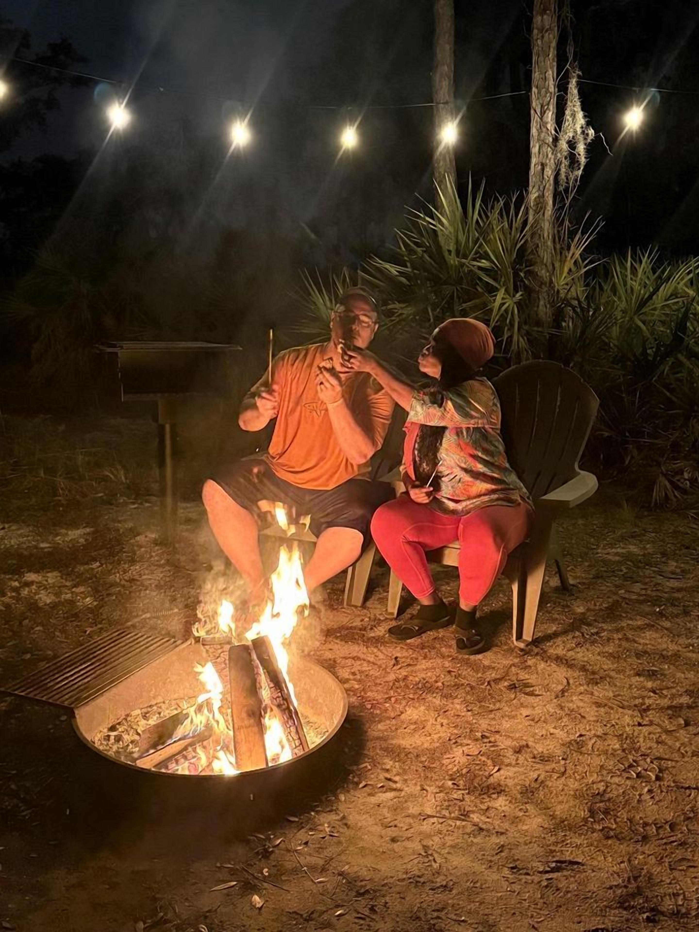 Deux personnes assises sur des chaises, autour d’un feu de camp, la nuit.