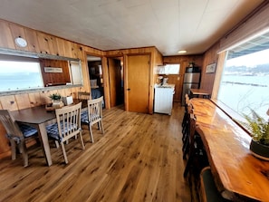 Un interior de madera con comedor, cocina y ventanas amplias con vista a un cuerpo de agua.
