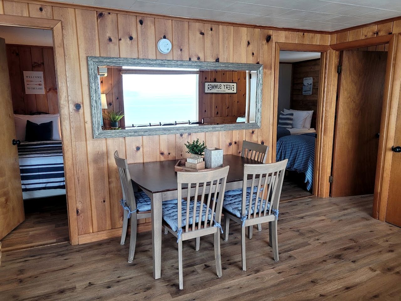 A wooden interior with a dining table and chairs, a mirror, and a sign reading 'WELCOME TO THE LAKE.'
