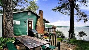 Uma cabana à beira do lago com mesa e cadeiras de piquenique, chaminé de tijolos e vista para o lago.