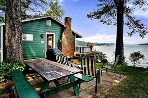 Eine Holzhütte am Seeufer mit Picknicktisch und -stühlen, einem Backsteinsschornstein und Blick auf den See.