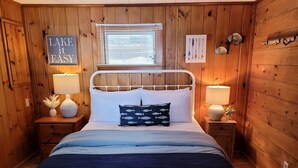Quarto de cabana de madeira com uma cama, duas mesas de cabeceira, uma janela com persianas e um letreiro que diz 'LAKE IT EASY'.