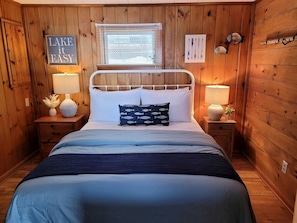 Un dormitorio de cabaña de madera con una cama, dos mesitas de noche, una ventana con persianas y un letrero que dice 'LAKE IT EASY'.