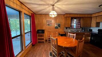 A wooden interior with a dining table, chairs, a kitchen area, and large windows with curtains.