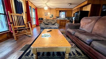 Una sala de estar con una mesa de centro de madera, un sofá de cuero, un tapiz estampado, sillas de madera y una cocina al fondo.