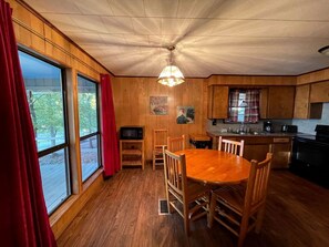 Un intérieur en bois comprenant une table à manger, des chaises, un coin cuisine et de grandes fenêtres avec des rideaux.