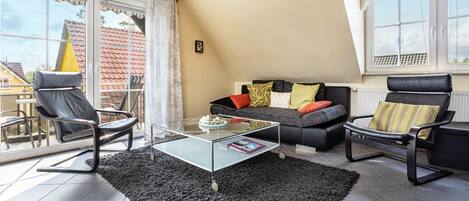 Una sala moderna con una mesa de centro de vidrio, dos sillones de cuero negro, un sofá negro con cojines coloridos y vista a un tejado rojo a través de amplios ventanales.