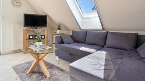 A modern living room with a sectional sofa, a glass coffee table, a flat-screen TV on a stand, a potted plant, and a skylight. - Apartment 'Haus 7 OG' with Lake View, Private Garden and Wi-Fi (Röbel)