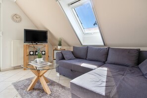 A modern living room with a sectional sofa, a glass coffee table, a flat-screen TV on a stand, a potted plant, and a skylight. - Apartment 'Haus 7 OG' with Lake View, Private Garden and Wi-Fi (Röbel)