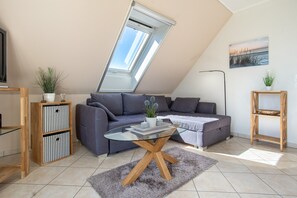 A modern living room with a sloped ceiling, a glass coffee table, a grey sofa, a wooden bookshelf, and a painting on the wall. - Apartment 'Haus 7 OG' with Lake View, Private Garden and Wi-Fi (Röbel)
