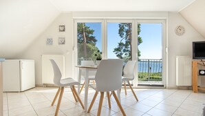 A modern kitchen with white appliances, a dining area with white chairs, and a large sliding glass door leading to a balcony with a view of trees and a body of water. - Apartment 'Haus 7 OG' with Lake View, Private Garden and Wi-Fi (Röbel)