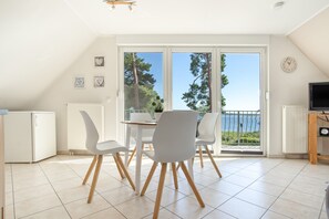 A modern kitchen with white appliances, a dining area with white chairs, and a large sliding glass door leading to a balcony with a view of trees and a body of water. - Apartment 'Haus 7 OG' with Lake View, Private Garden and Wi-Fi (Röbel)