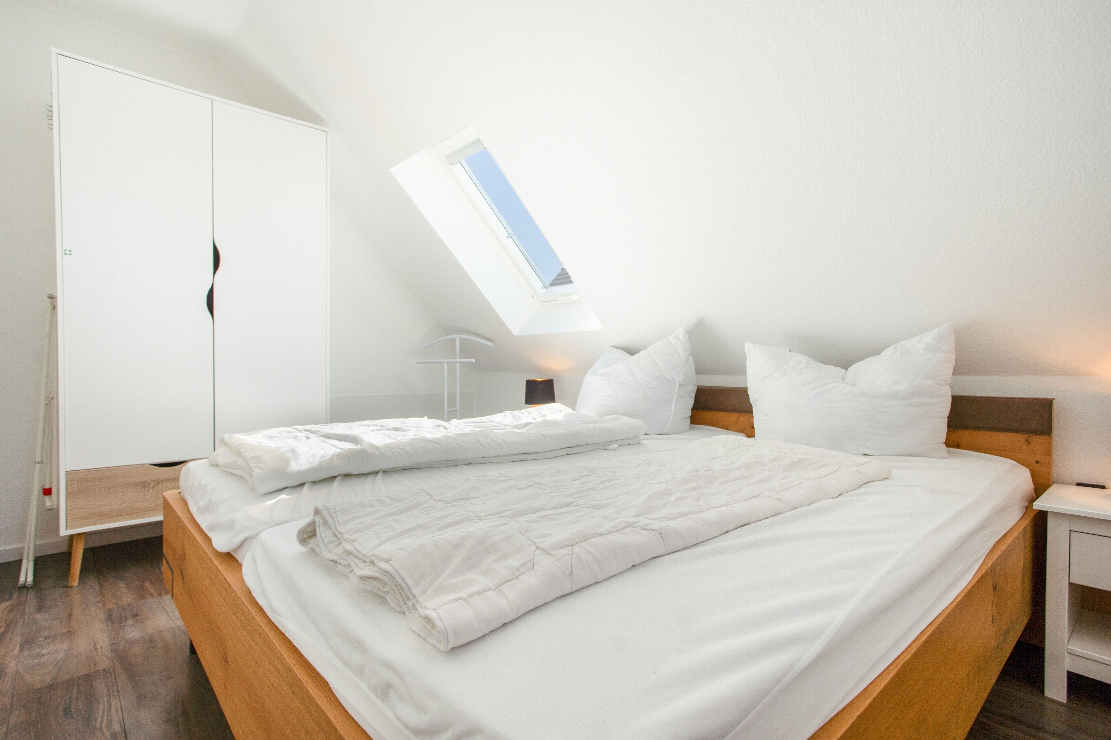 A neatly made bed with white bedding in a room with a skylight, a wardrobe, and a bedside table.