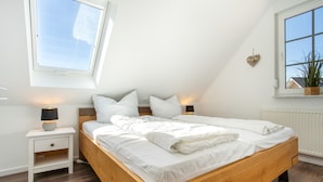 A bedroom with a wooden bed, white bedding, a nightstand with a lamp, a window with a view, and a plant on the windowsill. - Apartment 'Haus 7 OG' with Lake View, Private Garden and Wi-Fi (Röbel)
