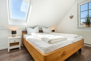 A bedroom with a wooden bed, white bedding, a nightstand with a lamp, a window with a view, and a plant on the windowsill. - Apartment 'Haus 7 OG' with Lake View, Private Garden and Wi-Fi (Röbel)