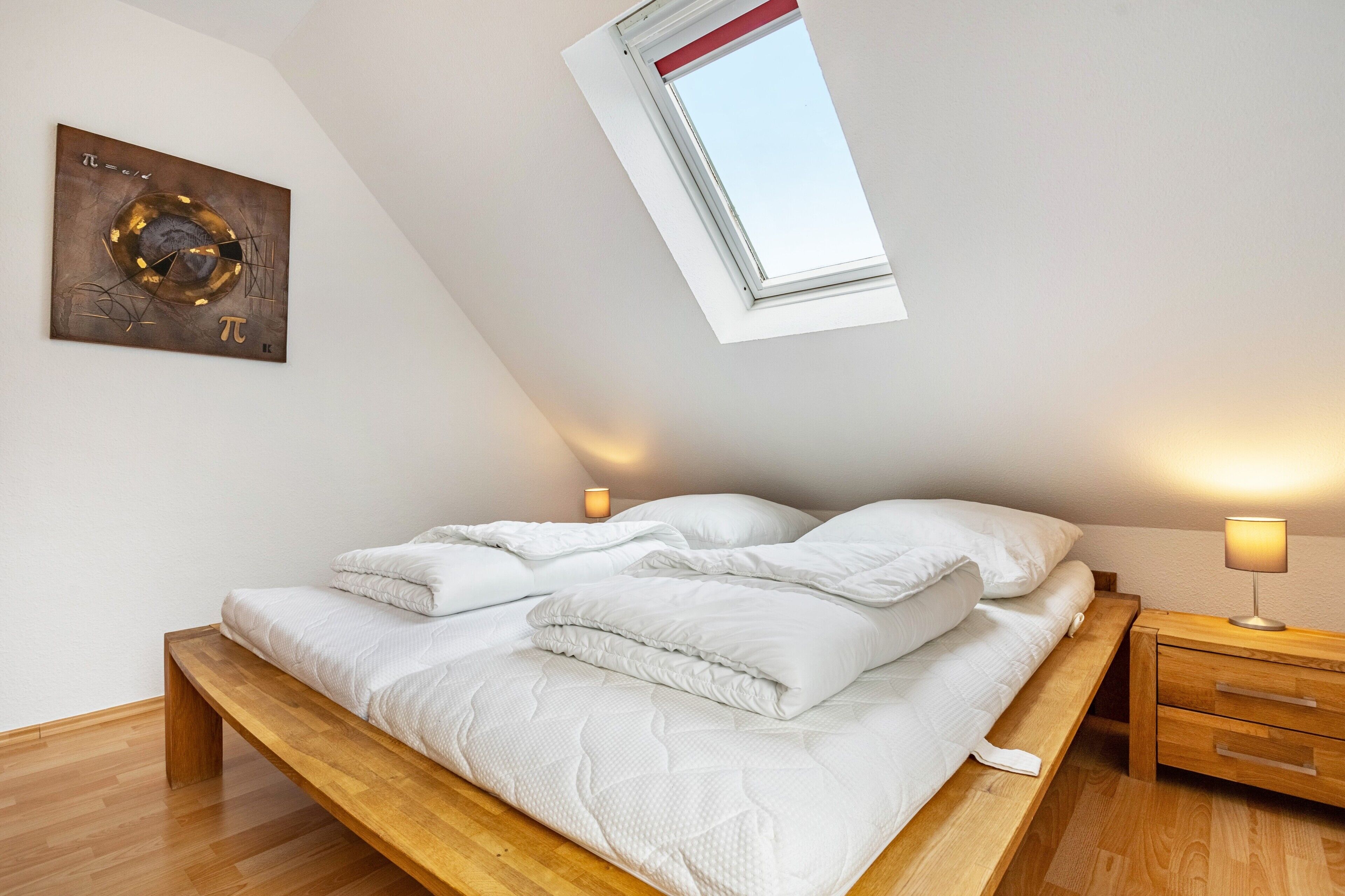 A bedroom with a sloped ceiling, a skylight, a bed with white bedding, a wooden bench, a bedside table with a lamp, and a wall clock.