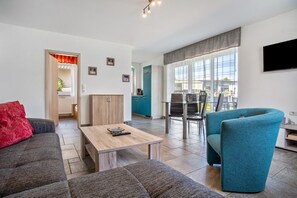 A modern living room with a sofa, a coffee table, and a teal armchair. There is a kitchen area with a blue cabinet and a dining area with a table and chairs. - Apartment 'Haus U13 EG' with Private Garden, Balcony and Wi-Fi (Röbel)