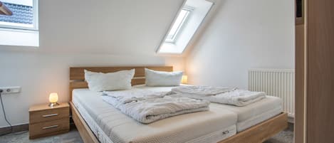 A bedroom with a sloped ceiling, a bed with white bedding, a bedside table with a lamp, and a window with a view of the roof.