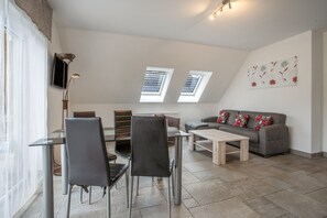 Una sala moderna con comedor, dos tragaluces, un sofá gris con almohadas rojas y un televisor de pantalla plana montado en la pared.