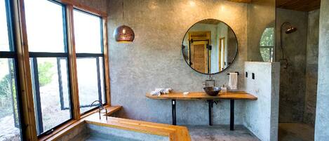 A modern bathroom with a large window, a round mirror, a wooden vanity, and a glass-enclosed shower area.