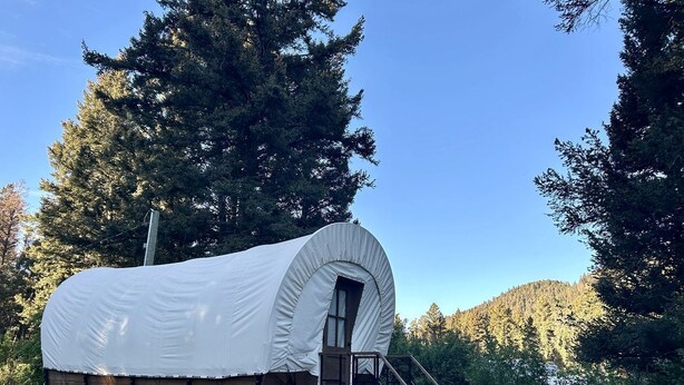A covered wagon with a small porch and steps leading up to it, set in a forested area. - Rustic Conestoga Wagon Stay with Lodge Amenities near Boulder Creek, Montana (Hall)