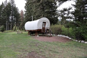 Eine blockhaüschenartige Hütte im Stil eines Wagen mit einer Holztreppe, die zu einer Tür führt, umgeben von einem Kiesweg und Grünpflanzen.