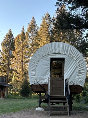 A covered structure with a door and steps leading up to it, surrounded by trees. - Rustic Conestoga Wagon Stay with Lodge Amenities near Boulder Creek, Montana (Hall)