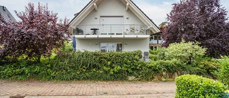 Ein zweistöckiges Wohngebäude mit Balkon, umgeben von Grünflächen und einer gepflasterten Auffahrt.
