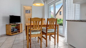 Uma área de refeições com mesa e cadeiras, uma televisão sobre um suporte, e uma porta de vidro deslizante que dá para o exterior.