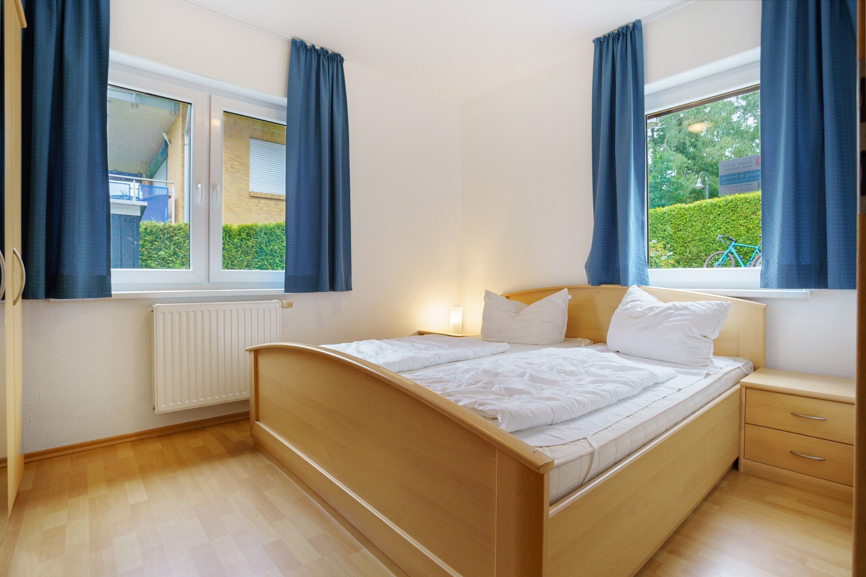 Une chambre à coucher avec un lit en bois, une table de chevet, une fenêtre avec des rideaux et un radiateur.
