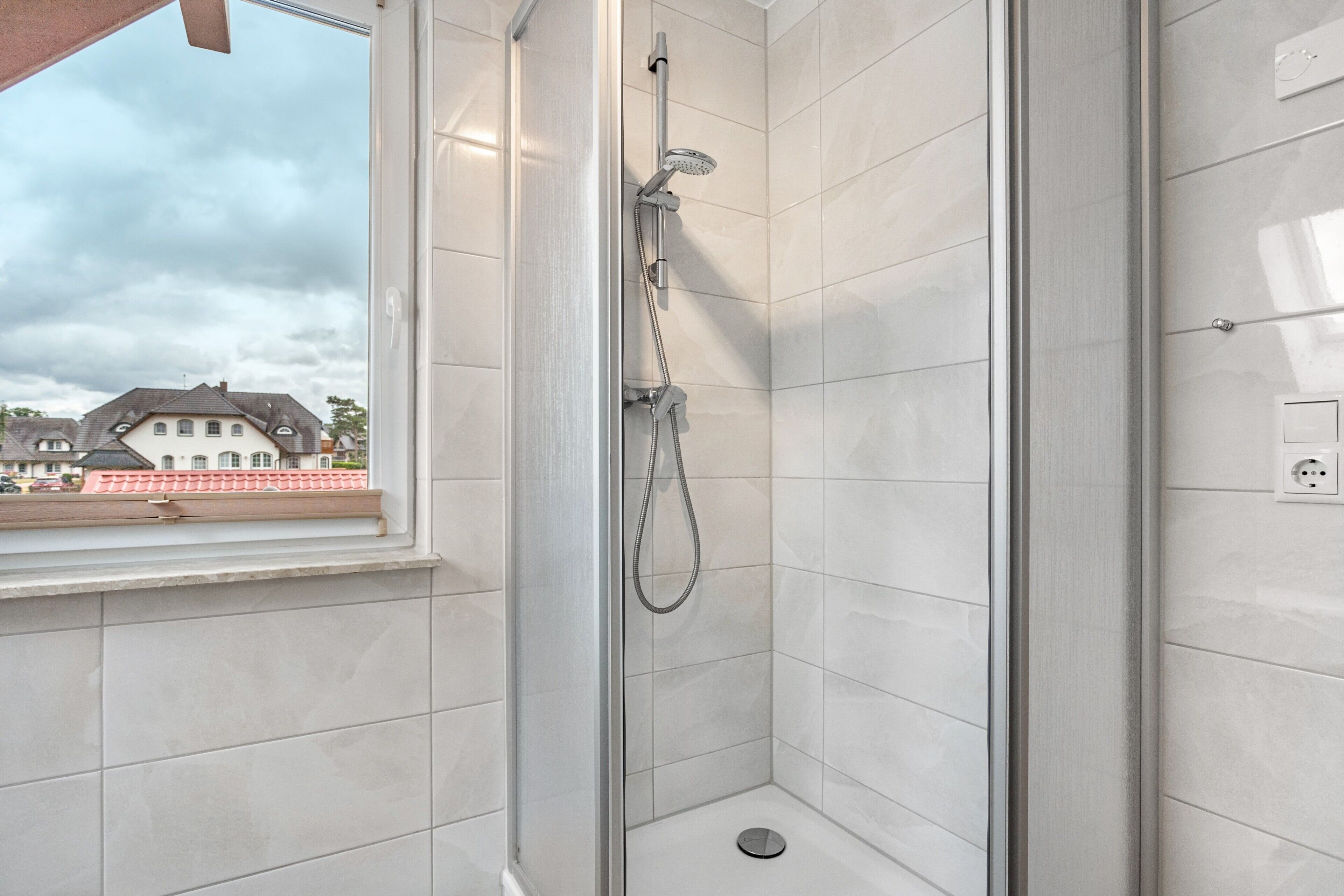 Ducha con puerta de vidrio, desagüe visible y ventana con vista a casas.