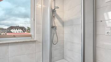Ducha con puerta de vidrio, desagüe visible y ventana con vista a casas.