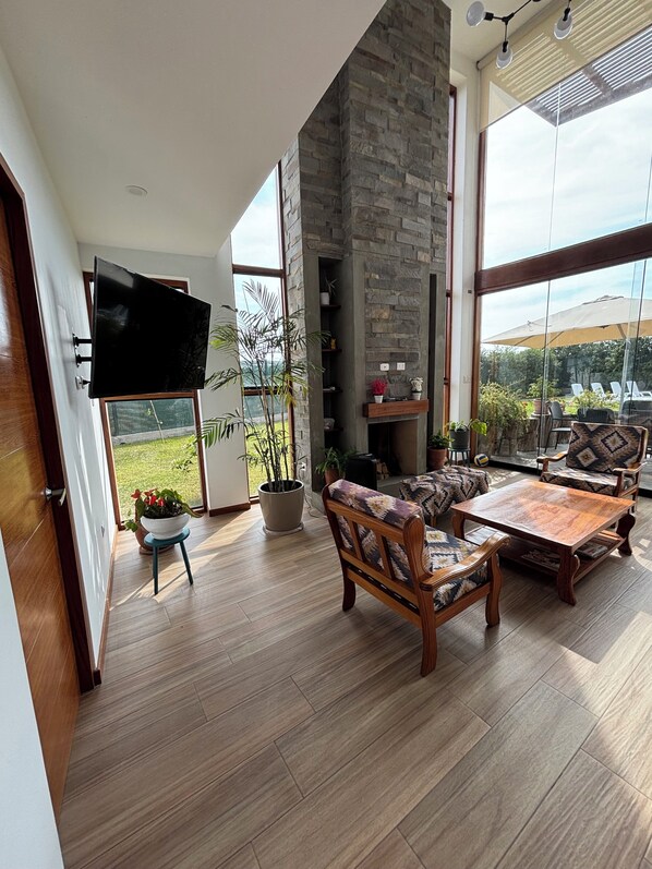 A spacious living room with a flat-screen TV, wooden furniture, a stone fireplace, and large windows offering a view of the outdoors.