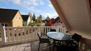 Una terraza de madera con mesa y sillas, barandal blanco y casas al fondo.