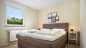 Een slaapkamer met een groot bed, een nachtkastje met een lamp, een radiator en een raam met uitzicht op groen.