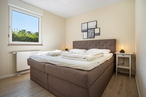 Ein Schlafzimmer mit einem großen Bett, einem Nachttisch mit Lampe, einem Heizkörper und einem Fenster mit Blick auf Grünflächen.