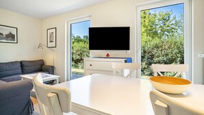 Een moderne woonkamer met een bank, een tafel, stoelen, een flatscreen televisie en een groot raam met uitzicht op groen.