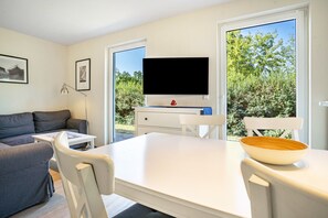Ein modernes Wohnzimmer mit einer Couch, einem Tisch, Stühlen, einem Flachbildfernseher und einem großen Fenster mit Blick auf Grünflächen.