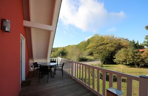 Ein Balkon mit Tisch und Stühlen, einem weißen Geländer und Blick auf Bäume sowie ein Gebäude in der Ferne.