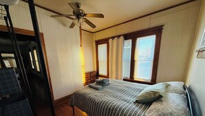 Un dormitorio con cama, ventilador de techo, ventana con persianas, mesita de noche y una silla.