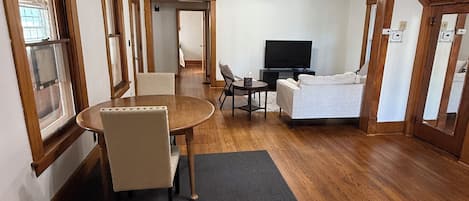 A spacious living room with a wooden floor, a dining table with chairs, a television, and a ceiling fan.