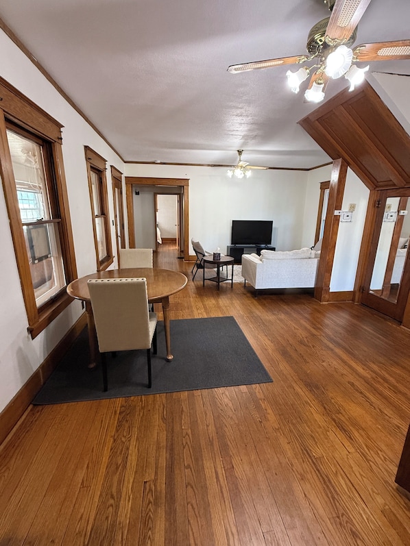 A spacious living room with a wooden floor, a dining table with chairs, a television, and a ceiling fan.