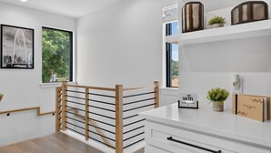 A modern hotel room with a white color scheme, a wooden staircase, a desk with a plant, and a view of greenery outside. - Luxury Townhome A. Walk to AT&T Stadium. Rooftop Terrace + Game Room. Sleeps 10. (Arlington)