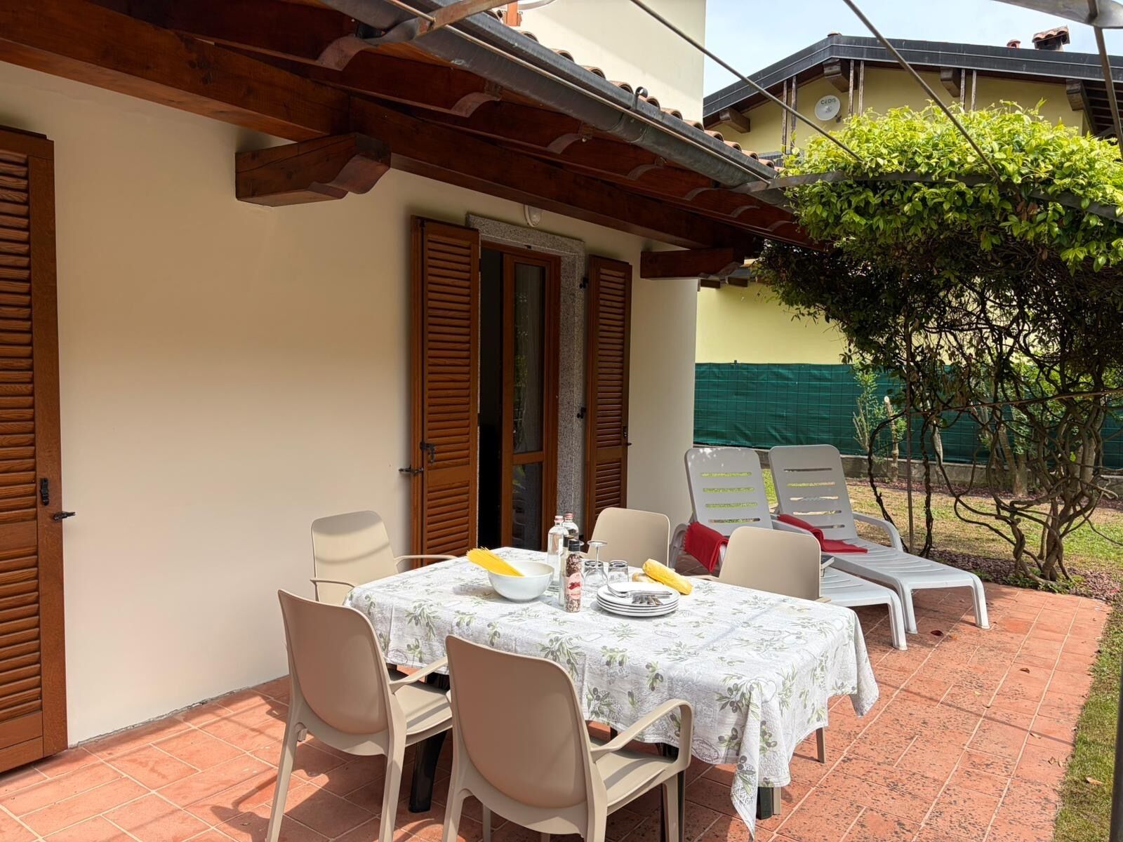 A patio with a table set for two, surrounded by chairs, and a small garden area with a tree and a fence.