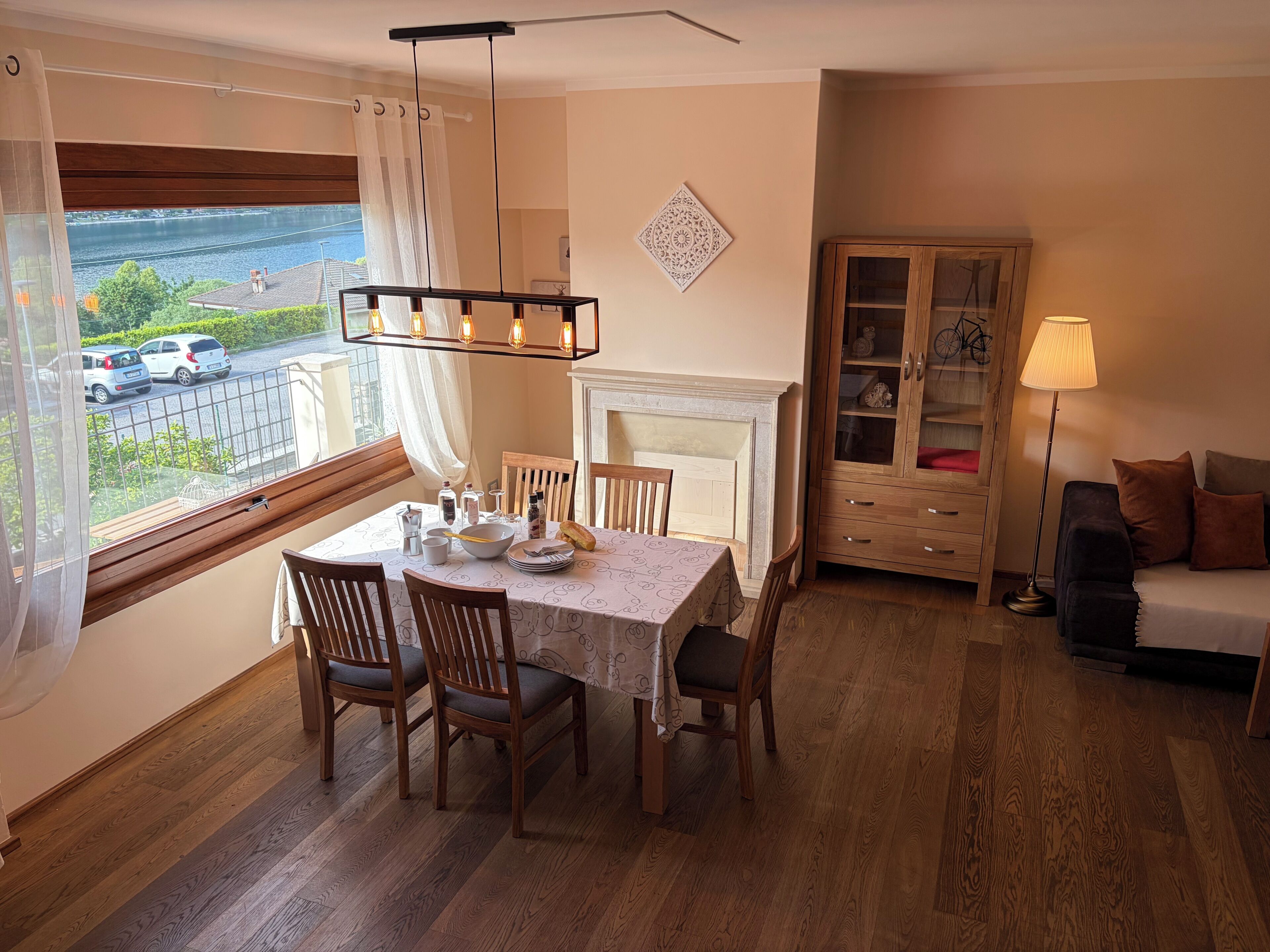 A dining area with a table set for four, wooden chairs, a chandelier, a view of a river, a cabinet with glass doors, and a sofa.