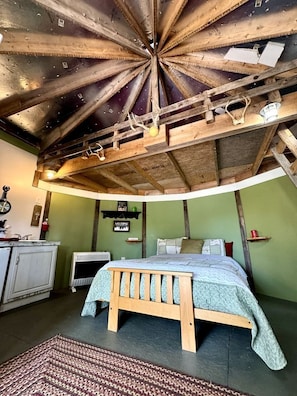 A room with a wooden ceiling, a bed, a radiator, a small shelf, and a welcome sign.