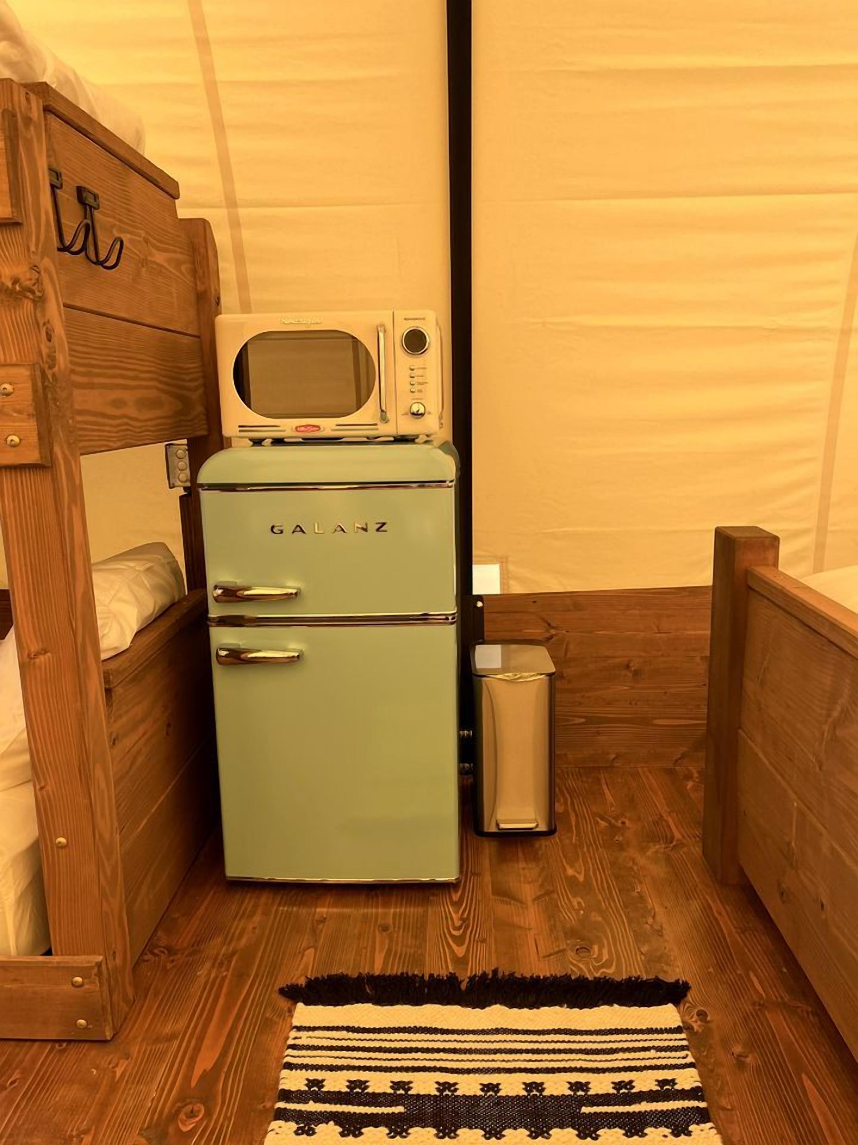 Una habitación pequeña con un refrigerador Galanz antiguo, una cama nido de madera, un cubo de basura pequeño y una alfombra estampada en el suelo.