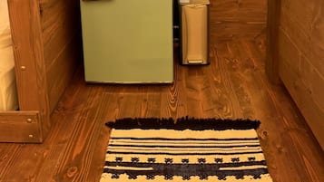 Una habitación pequeña con un refrigerador Galanz antiguo, una cama nido de madera, un cubo de basura pequeño y una alfombra estampada en el suelo.