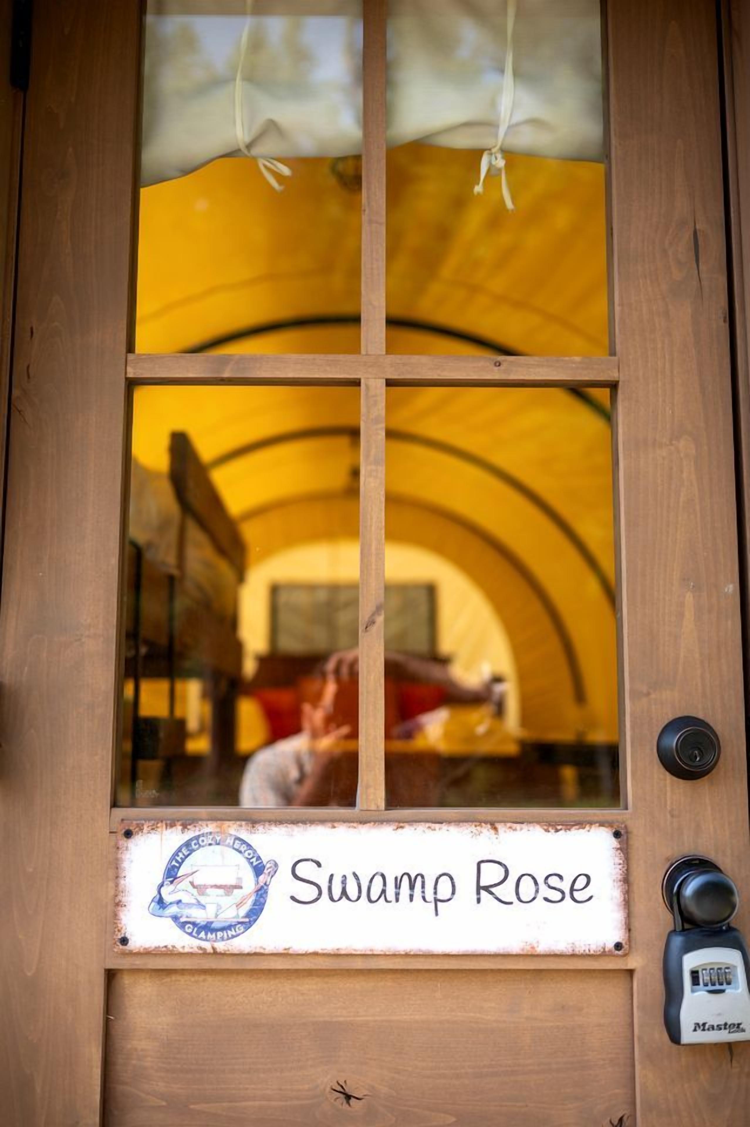 A wooden door with a sign reading 'Swamp Rose' and a view of a yellow tented area inside.