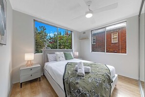 Ein modernes Schlafzimmer mit einem großen Bett, einem Nachttisch, einem Fenster mit Blick auf Bäume und einem Deckenventilator.